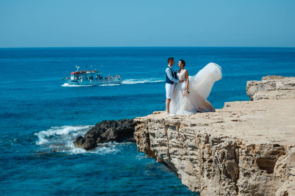schöne lächelnde junge Braut und Bräutigam, die am Strand spazieren gehen, sich küssen und Spaß haben, Hochzeitszeremonie in der Nähe von Felsen und Meer. Hochzeitszeremonie an der Küste Zyperns - Foto, Bild