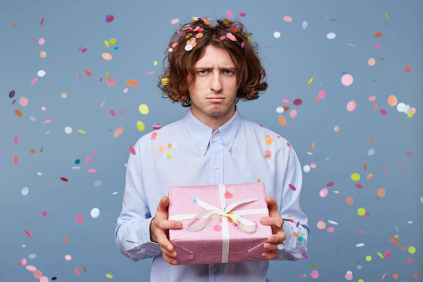 Porträt eines jungen Mannes, der eine geschmückte rosa Schachtel mit Geschenk in der Hand hält, sieht traurig, aufgebracht, frustriert, unzufrieden über etwas aus, das vor dem Hintergrund fallender Konfetti steht. - Foto, Bild