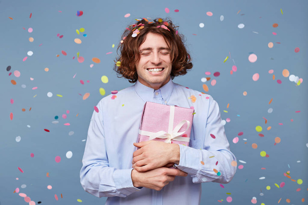 Birthday boy with eyes closed, presses the gift box tied with a white ribbon to himself,is grateful happy, enjoys moment, confetti falls on him,stands isolated on a blue background, feels happiness - Foto, afbeelding