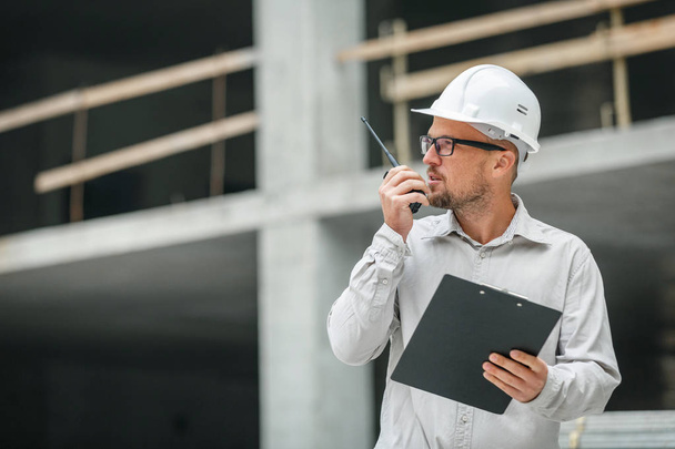 Male head engineer wearing white safety hardhat with walkie talkie and clipboard inspecting construction site. Development and construction industry concept - Photo, image