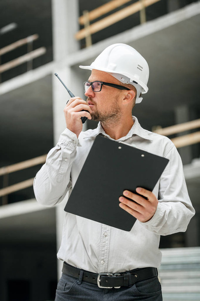 Male head engineer wearing white safety hardhat with walkie talkie and clipboard inspecting construction site. Development and construction industry concept - Foto, immagini