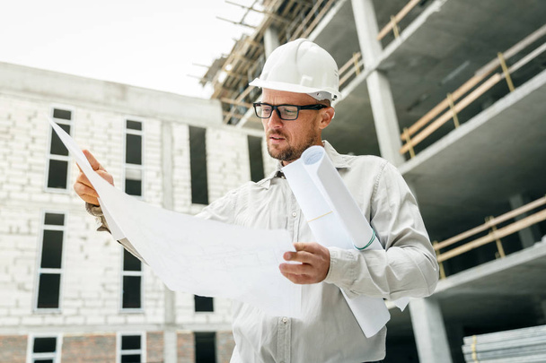 Construction engineer in a white hardhat inspecting blueprints on a construction site. Development and construction industry concept - Foto, immagini