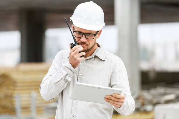 Male head engineer wearing white safety hardhat with walkie talkie and tablet inspecting construction site. Technology, development and construction industry concept - Photo, image