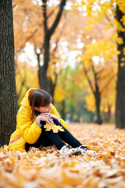 Sad teen girl sits near tree in autumn park. Bright yellow leaves and trees. - Foto, imagen
