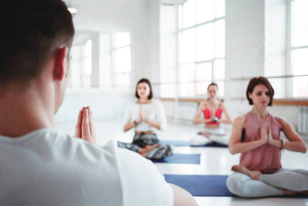 Man coach trains group of women yoga exercises for maintaining health care in white class. People together practis yoga poses. Blurred background - 写真・画像