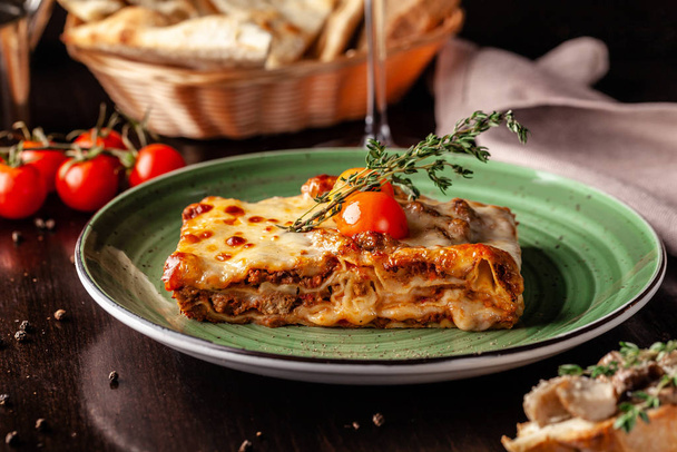 The concept of Italian cuisine. Baked lasagna with minced bolognese, pasta, cherry tomatoes lies on a green plate in a restaurant. A glass of red wine and bread basket on the table - Zdjęcie, obraz