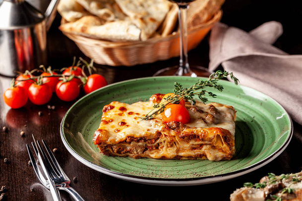 The concept of Italian cuisine. Baked lasagna with minced bolognese, pasta, cherry tomatoes lies on a green plate in a restaurant. A glass of red wine and bread basket on the table - Zdjęcie, obraz