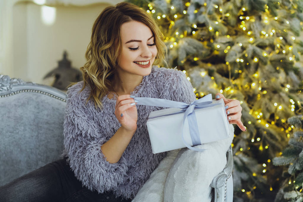 Young beautiful blonde woman in a gray sweater with a gift on hands near the New Year, Christmas tree with gifts - 写真・画像