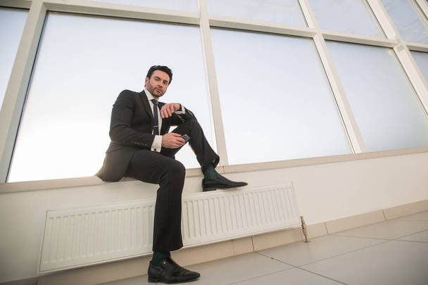 confident businessman with a smartphone sitting in the office hallway - Zdjęcie, obraz