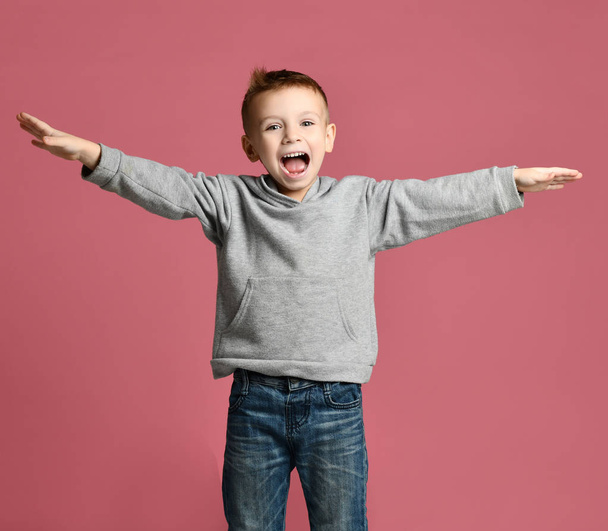 Young boy kid jumping in grey hoodie with hands spread up laughing smiling on pink  - Φωτογραφία, εικόνα