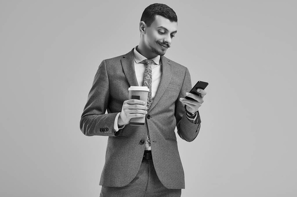 ファッション灰色完全なスーツに派手な口髭のハンサムな若い自信アラビア語ビジネスマンの肖像画を保持する一杯のコーヒー、スタジオの背景に電話でメッセージを入力. - 写真・画像