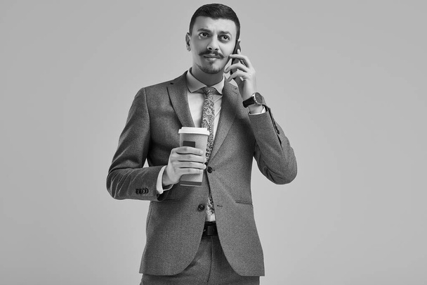 Porträt eines hübschen jungen selbstbewussten arabischen Geschäftsmannes mit schickem Schnurrbart im modischen grauen Ganzanzug hält eine Tasse Kaffee in der Hand und telefoniert - Foto, Bild