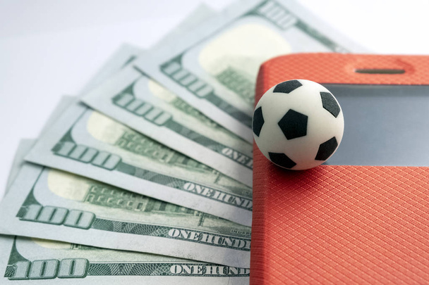 A football souvenir ball on the corner of a mobile phone screen in a red case against a background of five hundred US dollars. The concept of sports betting with a smartphone. - Photo, Image