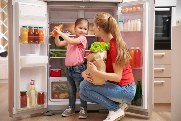 Молода мама з дочкою кладе їжу в холодильник вдома після покупки
 - Фото, зображення