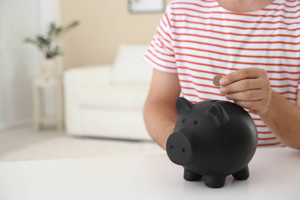 Man putting coin into piggy bank on table against blurred background, closeup with space for text - Foto, Imagen