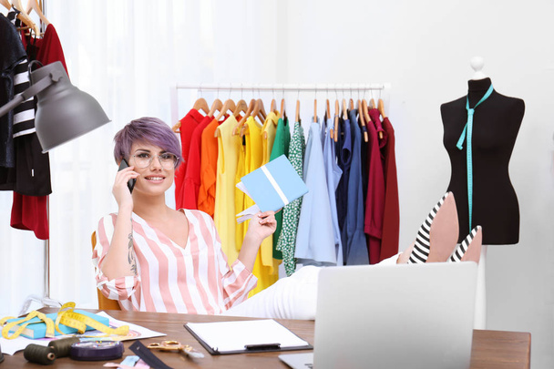 Beautiful young stylist talking on phone at workplace near rack with clothes - Photo, Image