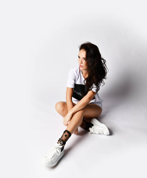 Beautiful brunette woman sitting on grey background in long white t-shirt and sneakers - Fotó, kép
