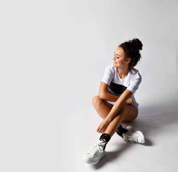 Beautiful brunette woman sitting on grey background in long white t-shirt and sneakers - Fotoğraf, Görsel