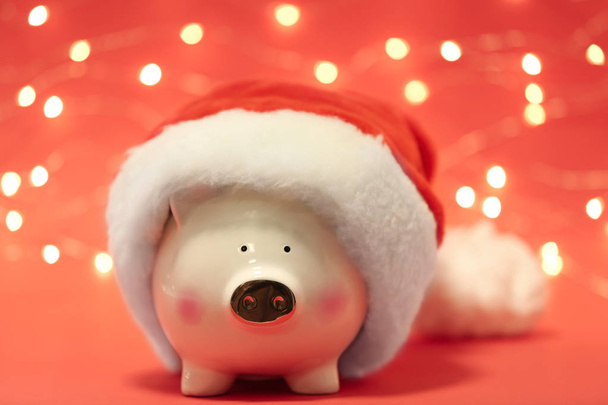 Cute piggy bank with Santa hat against blurred Christmas lights - Photo, image