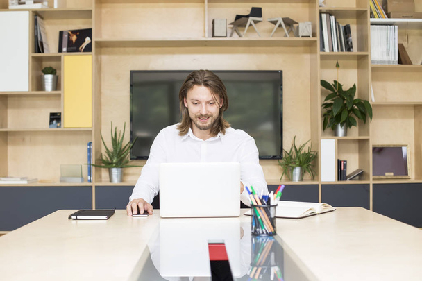 Portré egy alkalomszerűen öltözött fiatal üzletemberről, aki laptopon dolgozik, miközben egy íróasztalnál ül egy irodában. - Fotó, kép