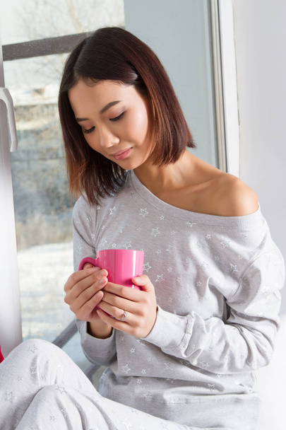 Chica en pijama sentada en el alféizar de la ventana con una taza en las manos
 - Foto, Imagen