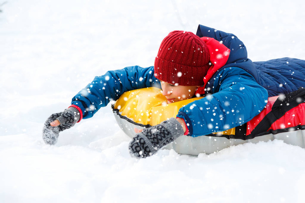 cute young boy in hat red scarf and blue jacket lays on tube on snow, has fun, smiles. Teenager rises his hands up in winter park. Active lifestyle, winter activity, outdoor winter games, snowballs - Photo, Image