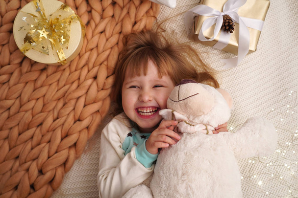 メリノ ベージュからデザイナーの毛布。笑う赤ちゃんがベッドで横になっています。幸せな女の子は、新年の受け取ったテディベアを抱擁します。クリスマスの発症時喜びベッドに横になっている幸せな子供 - 写真・画像