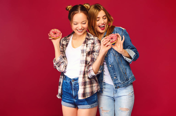 Twee mooie lachende hipster meisjes poseren in trendy zomer geruit overhemd kleding. Vrouwen geïsoleerd op rode achtergrond. Positieve modellen houden van verse roze donuts met poeder klaar om te genieten van snoep - Foto, afbeelding