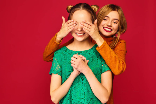 Photo of two beautiful blond ladies standing isolated over red background. Smiling girl covering eyes with hands to her friend. Positive woman models hugging  - Φωτογραφία, εικόνα