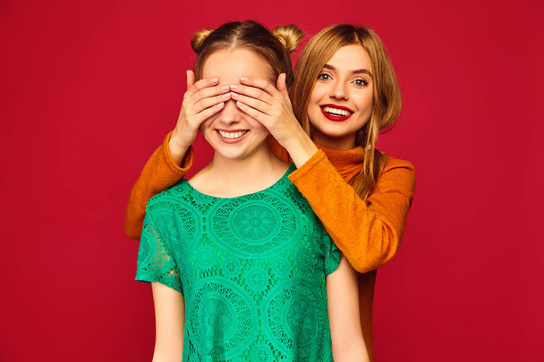 Photo of two beautiful blond ladies standing isolated over red background. Smiling girl covering eyes with hands to her friend. Positive woman models hugging  - Fotó, kép