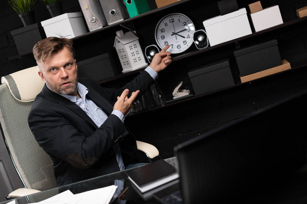 Empresario sentado en la mesa de la computadora y apunta sus manos a la esfera del reloj
 - Foto, Imagen