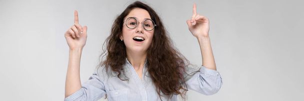 Young brunette girl with glasses. The girl pulls her lips for a kiss. The girl put her hand to her head. The girl points with her fingers up. - Photo, Image