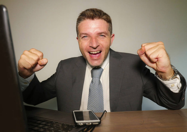 young happy and attractive business man working with mobile phone at office computer desk celebrating success gesturing excited in corporate company job success and executive lifestyle - Фото, зображення