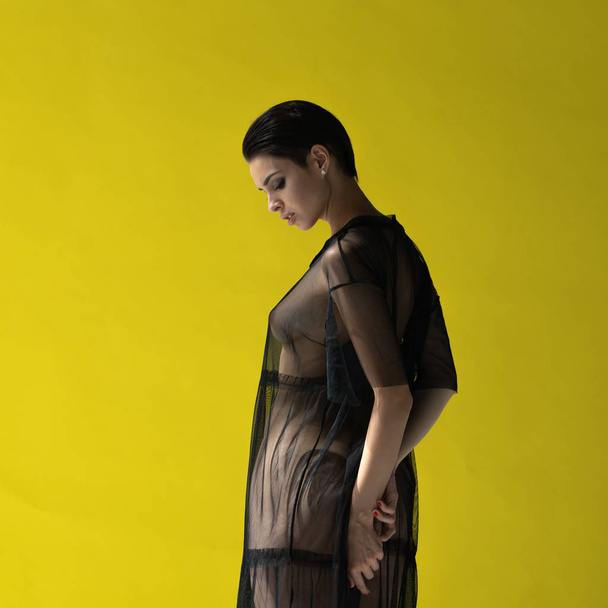 Młoda dziewczyna piękny pozowanie nago w studio, stojąc w czarna sukienka w pobliżu ściana żółty - Zdjęcie, obraz
