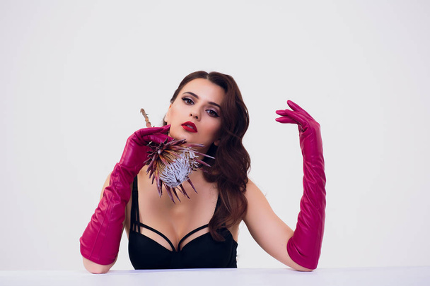 Beautiful woman holding protea flower in white room Retro red silk gloves, dark dress, woman with curly hair, brunette girl on white background - Φωτογραφία, εικόνα