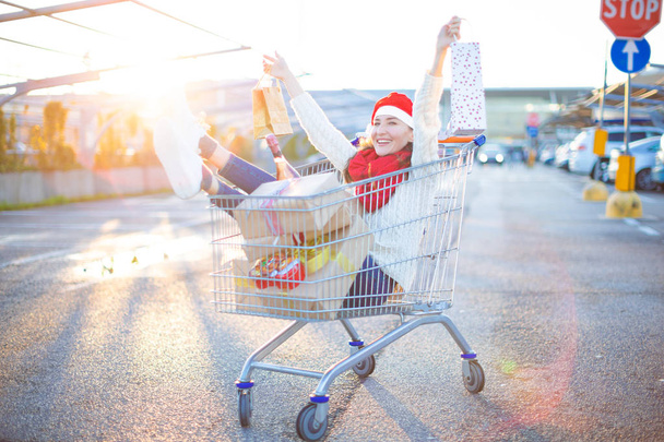 Happy young smiling woman sitting in shopping cart with a lot of gift boxes and two shopping bags in her hands wearing red Santa Christmas hat - holiday purchases and celebration concept. - Fotó, kép