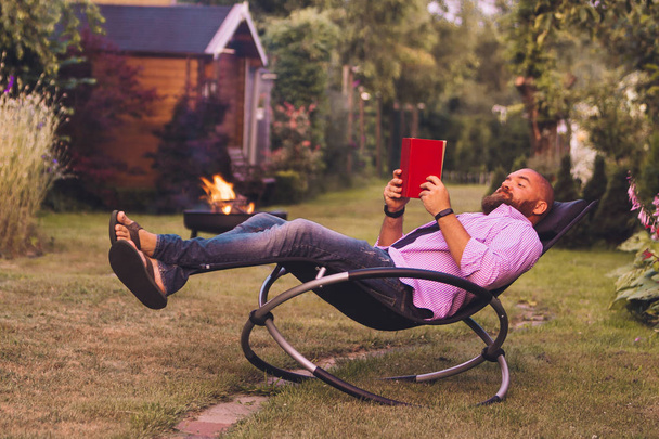 Bearded man reading book on a rocking chair (rocker) in the garden - Фото, изображение