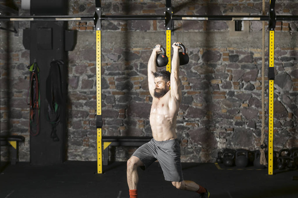 Young bearded and shirtless athlete warming up with kettlebells for workout at the gym.  Dark photography concept with copy space area.  - Foto, immagini