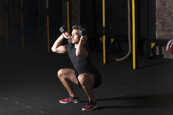 Young male athlete practicing dumbbell squats at the gym. Dark photography concept with copy space text area. - Photo, Image