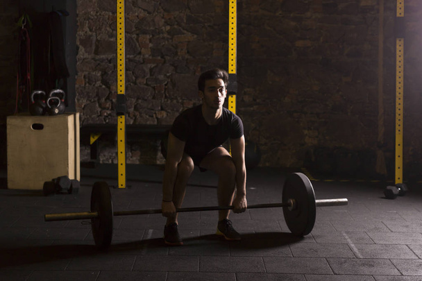 Young athlete training with a heavy barbell at the gym. Dark photography concept with copy space area. - Foto, Imagen