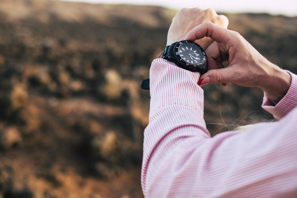 close up of woman hands checking and touching a black clock watch during an outdoor excursion in a desert place - rocks defocused background - control time concept - Photo, image
