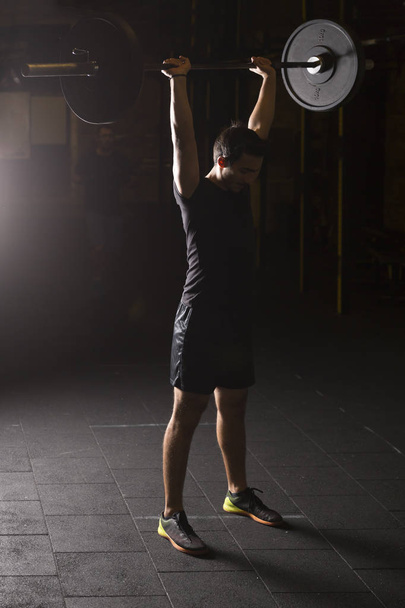 Молодой спортсмен тренируется с тяжелой штангой в спортзале. Концепция темной фотографии с зоной копирования
. - Фото, изображение
