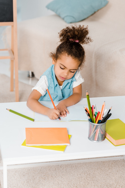 high angle view of cute african american child sitting and writing with pencil - Foto, imagen