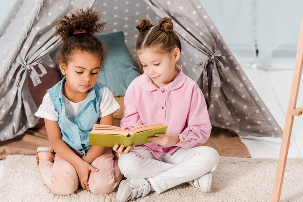 high angle view of cute multiethnic kids sitting on carpet and reading book together  - Photo, Image