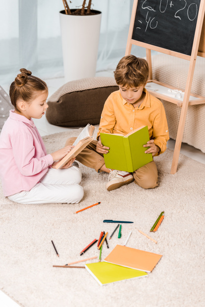 Blick von oben auf entzückende fokussierte Kinder, die auf Teppich sitzen und gemeinsam Bücher lesen    - Foto, Bild