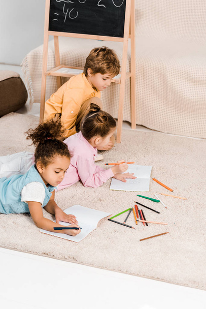 high angle view of cute multiethnic children lying on carpet and studying together  - 写真・画像