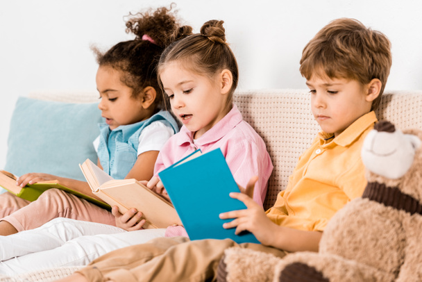 hermosos niños multiétnicos sentados en el sofá y leyendo libros juntos
 - Foto, imagen