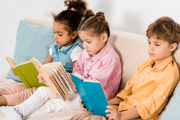 adorable multiethnic children sitting on sofa and reading books  - Fotó, kép