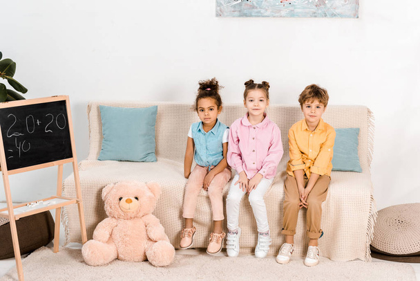 belas crianças multiétnicas sentadas no sofá e olhando para a câmera
  - Foto, Imagem