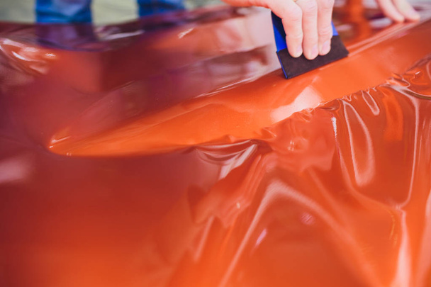 Car wrapping specialist putting vinyl foil or film car wrapping protective film yacht, boat, ship, car, mobile home. orange film heating with hair dryer and trimming plastic soft hard squeegee - Фото, изображение